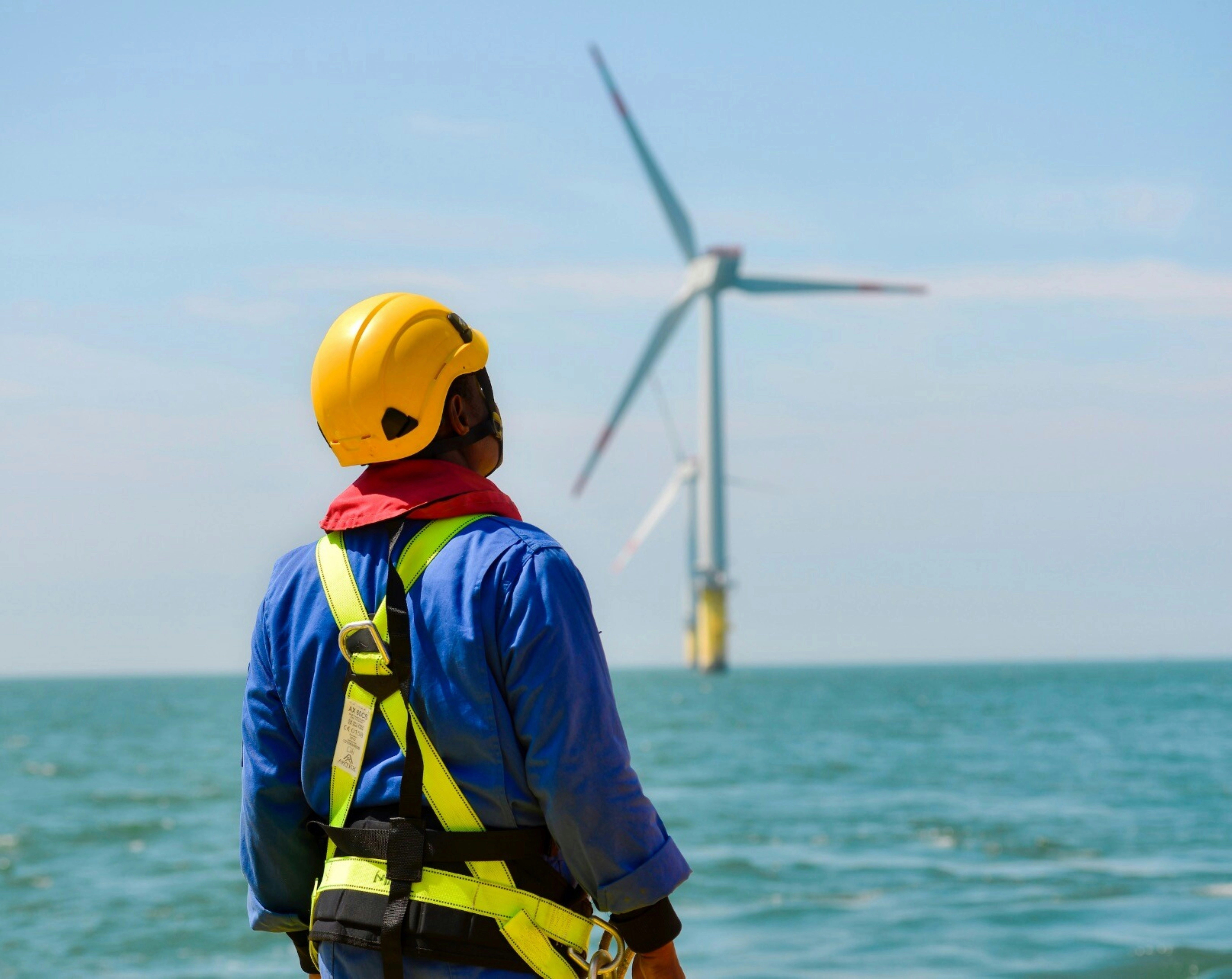 Technicien de maintenance éolien équipé d'un harnais et regardant une éolienne en mer